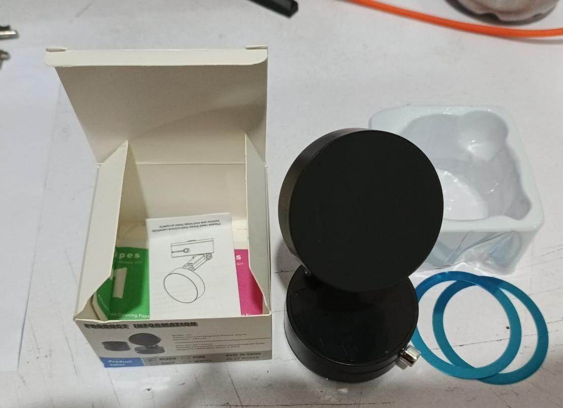 A round, black, magnetic phone holder next to its open white box, a white plastic tray, and two blue metallic rings.